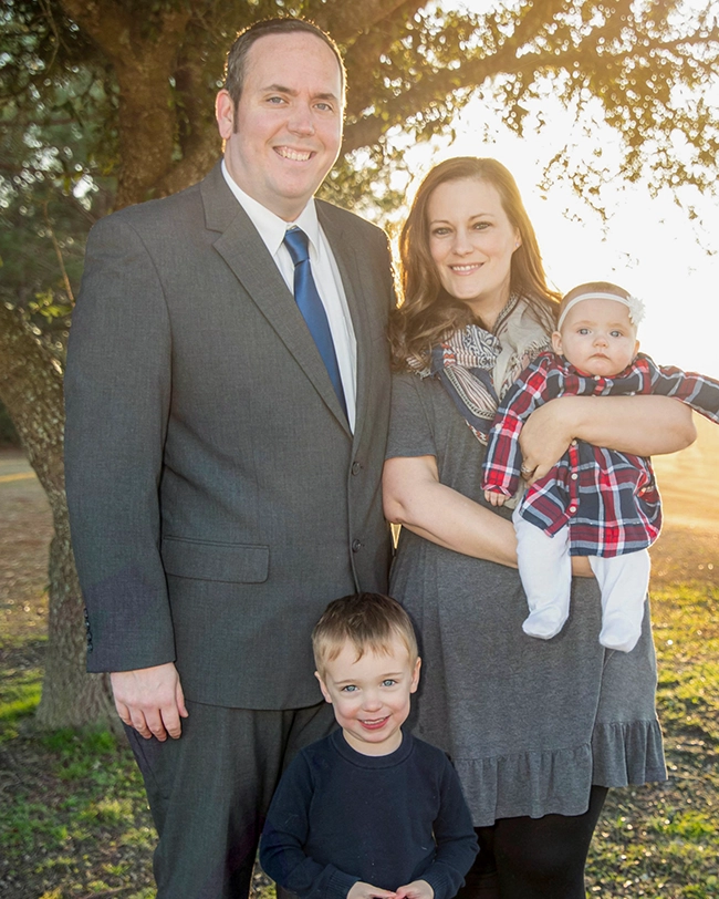 Chris Schulte in a suit and tie standing near a tree with his wife and children.